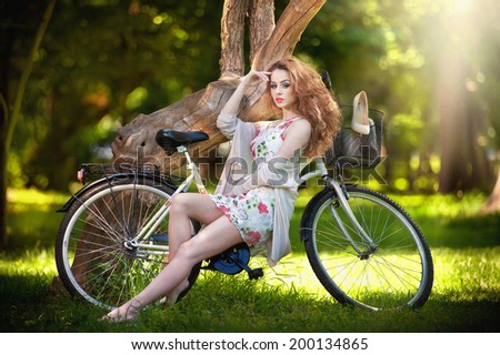 Beautiful girl wearing a nice short dress having fun in park with bicycle. Pretty red hair woman with romantic look posing sitting on her bike in a sunny day. Gorgeous curly redhead relaxing outdoor.