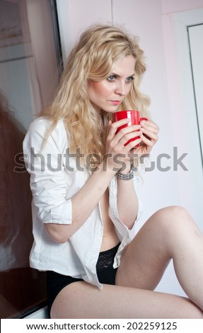 portrait of a young, blond woman, holding a mug with both her hands, wearing a white shirt and black pants, with an expression of being sadness. Woman posing with a big red cup of tea. in  her hands.