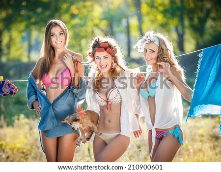 Three sexy women with provocative outfits putting clothes to dry in sun. Sensual young females laughing putting out the washing to dry in sunny day. Perfect body housewives with a dog, shot in forest