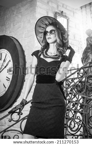 Young beautiful brunette woman in black standing on stairs near an over sized wall clock. Elegant romantic mysterious lady with movie star look in luxurious vintage interior, black and white photo.