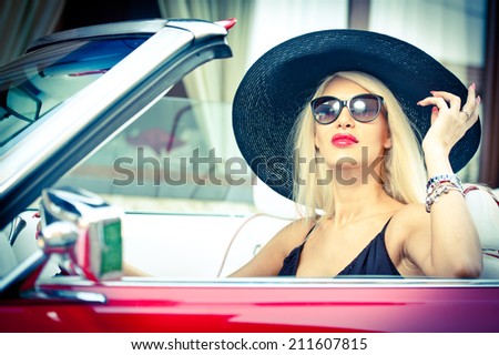 Outdoor summer portrait of stylish blonde vintage woman driving a convertible red retro car. Fashionable attractive fair hair female with black hat in red vehicle. Sunny bright colors, outdoors shot.