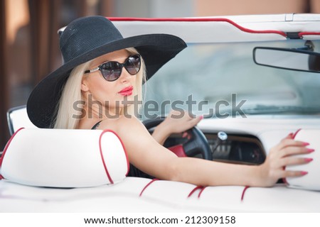 Outdoor summer portrait of stylish blonde vintage woman driving a convertible red retro car. Fashionable attractive fair hair female with black hat in red vehicle. Sunny bright colors, outdoors shot.