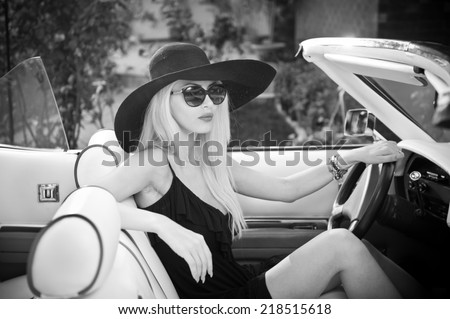 Outdoor summer portrait of stylish blonde vintage woman driving a convertible retro car. Fashionable attractive fair hair female with black hat in withe leather vehicle. Black and white outdoors shot.