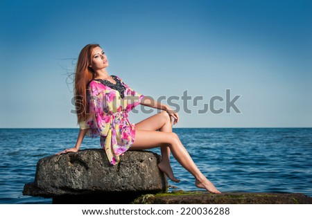 Fashion portrait of young woman with long red hair hair in a colored blouse at the beach. Sensual attractive girl on a rock near the sea. Woman with perfect body relaxing on the rock near the ocean