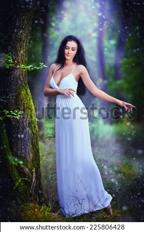 Lovely young lady wearing an elegant long white dress enjoying the beams of celestial light on her face in enchanted woods. Long hair brunette woman looking as a glamorous princess in the forest