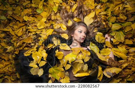 Nice girl covered with autumnal leaves. Young woman laying down on the ground covered by fall foliage in park. Beautiful sexy girl lying on autumn leaves. Young woman covered with golden autumn leaves