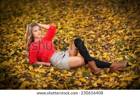 Beautiful elegant woman with red blouse and short skirt posing in park during fall. Young pretty woman with blonde hair lying down on autumnal leaves. Sensual blonde with black leggings in forest.