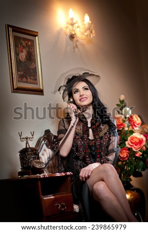 Young beautiful luxurious woman in long elegant dress talking by phone in a elegant scenery. Seductive brunette woman with roses in background holding a vintage phone in luxurious classic interior