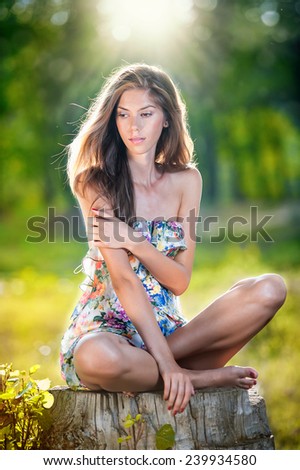 Young beautiful long hair woman wearing a multicolored dress posing on a stump in a green forest. Fashionable sexy attractive girl sitting on hub in a sunny day. Gorgeous girl in garden in summer day.