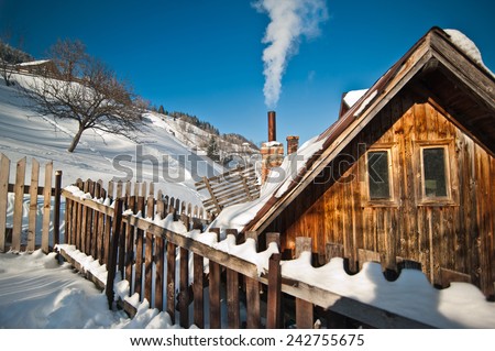 Old wooden cottage with hill covered by snow in background. Bright cold winter day in the mountains landscape. Carpathian mountains, Romania. Isolated wood mountain house cabin hut covered by snow