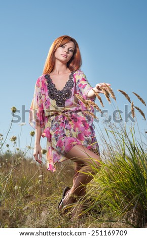 Beautiful young woman in wild flowers field on blue sky background. Portrait of attractive red hair girl with long hair relaxing in nature, outdoor shot. Lady in multicolored dress enjoying nature