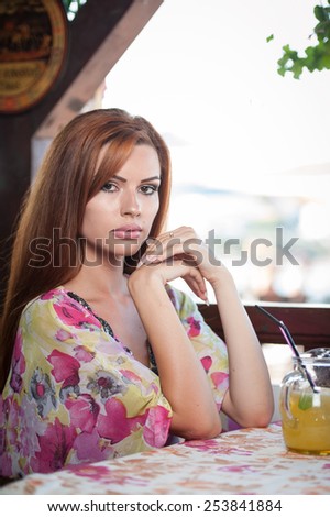 Attractive red hair young woman with bright colored blouse drinking lemonade on a terrace having blue sea in background. Gorgeous redhead model drinking fresh drink with straw in a summer day