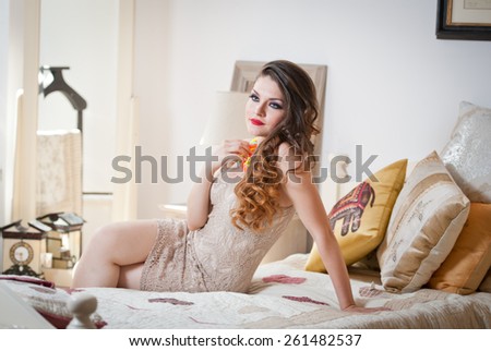 Young beautiful sexy woman in white short tight dress posing challenging indoor on vintage bed. Sensual long hair brunette in bedroom. Attractive female lying provocatively on bed full of pillows