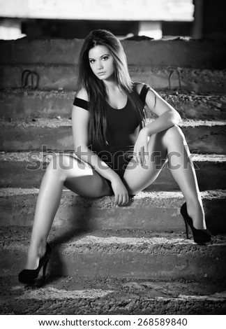 Fashionable pretty young woman with long legs sitting on old stone stairs. Beautiful long hair brunette on high heels shoes posing provocatively. Young model wearing black short dress, frontal view