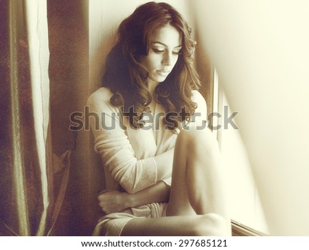 Attractive sexy brunette in white dress posing provocatively in window frame. Portrait of sensual woman in classic boudoir scene. Woman with long hair daydreaming and enjoying the bright day light
