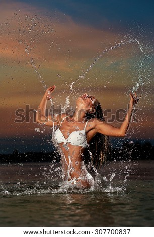 Sexy brunette woman in wet white swimsuit posing in river water with sunset sky on background. Young female playing with water in twilight scenery. Attractive girl in summer evening over dramatic sky.