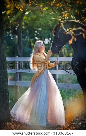 Fashionable lady with white bridal dress near black horse in forest. Beautiful young blonde woman in a long dress posing with a friendly horse. Attractive elegant female with horse, sunny summer day