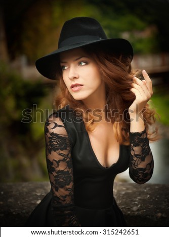 Charming young light brown hair brunette woman with big black hat and blouse with lace sleeves. Sexy gorgeous young woman with curly hair posing. Beautiful portrait of a sensual woman with long hair