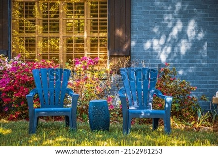 Two blue Adirondack chairs and matching drum table sitting on lawn in front of shuttered window in blue brick house landscaped with azaleas in dappled light Foto stock © 