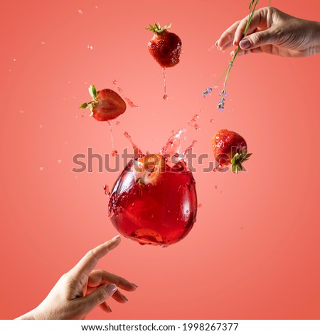 Woman hand support fly glass of strawberry drink with splash, juice strawberries falling in glass. Cocktail of strawberry and lavender flavor. Summer art food concept on pink background