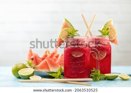 Watermelon mojito fresh red drink. With watermelon, lemon, lime, mint leaves and ice. The drink is on a white table with a cut watermelon in the background.