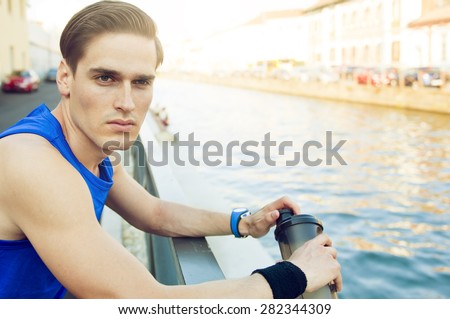 Picture of a young handsome athlete resting by the river on a sunny day after work out.He has protein shake at his hand.