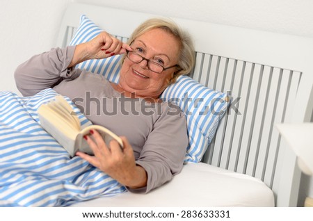 Happy Middle Aged Woman Wearing Eyeglasses, Holding a Novel Book While Lying on her Bed and Looking at the Camera.