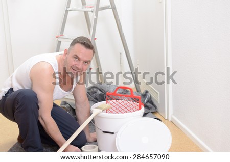 Muscular man doing DIY renovations kneeling down amidst tubs of paint , rollers, paintbrushes and a stepladder looking up at the camera with a smile