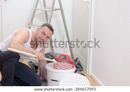 Muscular man doing DIY renovations kneeling down amidst tubs of paint , rollers, paintbrushes and a stepladder looking up at the camera