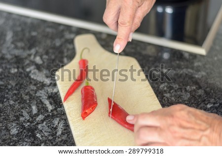 Elderly lady slicing red chili peppers with a kitchen knife on a wooden chopping board as she prepares a meal