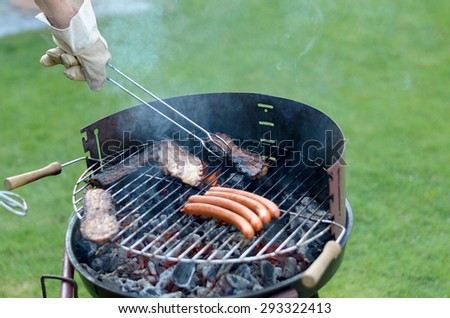Person wearing a glove using long handled tongs to grill spicy sausages over a portable barbecue outdoors on green grass at a summer picnic or campsite