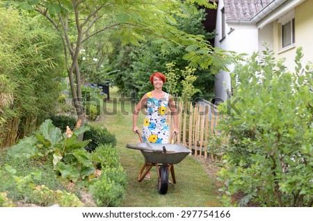 Happy Redhead Woman in Floral Dress, Pushing a Garden Wheelbarrow with Tools While Looking at the Camera.