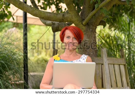 Adult Redhead Woman with Laptop Computer, Sitting on the Wooden Bench Under the Tree and Smiling at the Camera