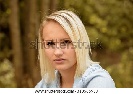 Pretty blond woman with a trendy haircut looking at the camera with a serious expression, head and shoulders portrait outdoors against trees