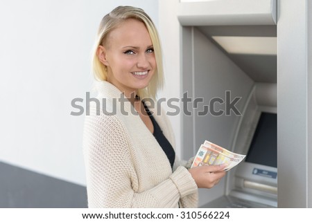Stylish woman drawing money at an outdoor bank ATM holding a handful of banknotes and turning to give the camera a lovely smile