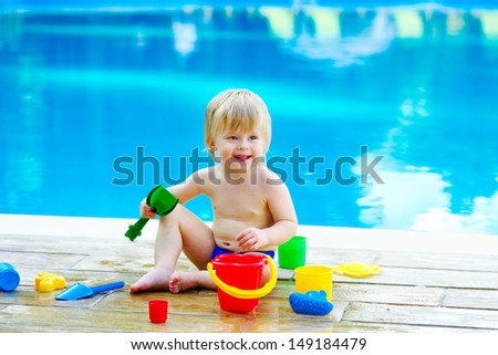 Adorable toddler playing with toy bucket and spades set by the swimming pool with blue water