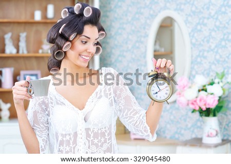 Morning rush. Beautiful woman in hair curlers holding cup of coffee and looking at the clock
