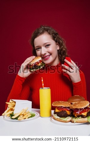 A curly young woman with bright pink lipstick on her lips smiling and looking at the camera, holding a cheeseburger in one hand and a red napkin in another hand over a vinous background.  Foto d'archivio © 