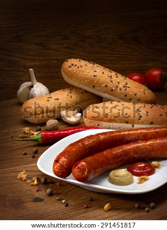 Sausages frying on a plate with sauce, bread rolls and condiments.
