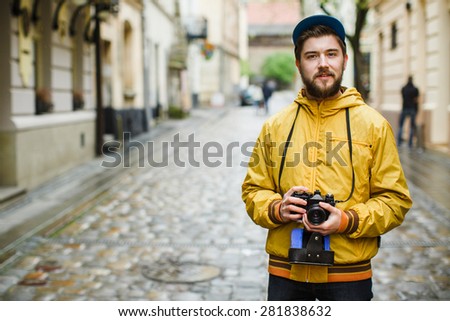 Young hipster with a beard holding oldshool film camera and looking at camera, on the street, wearing in bright raincoat and hat, portrait, day outdoors, ready to take a picture