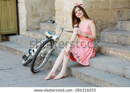 Young pretty blond-brown girl sitting on concrete stairs with a cup of coffee and looking at camera, with vintage bicycle, in an old European city