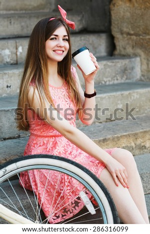 Young pretty blond-brown girl sitting on concrete stairs and smiling with a cup of coffee, looking at camera
