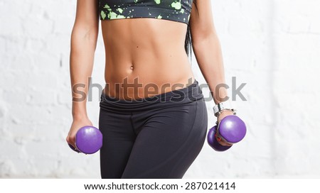 Athletic young woman showing some strong abs and flat belly, in sports outfits with beautiful body, holding purple dumbbells, against concrete wall