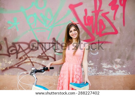 Pretty young girl with long blond-brown hair, in pink head wrap and dress with a pattern of flowers. Holding vintage bicycle on urban graffiti background.