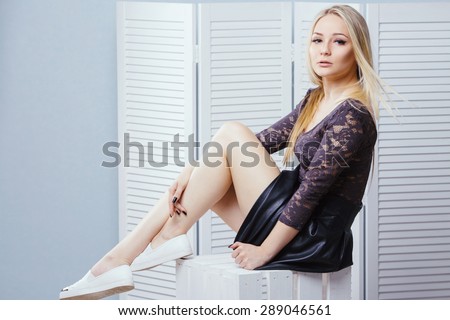 Sensual blonde young girl in a black skirt, lace top and white shoes sitting on white wooden box. On a white paravent background. Copy space.