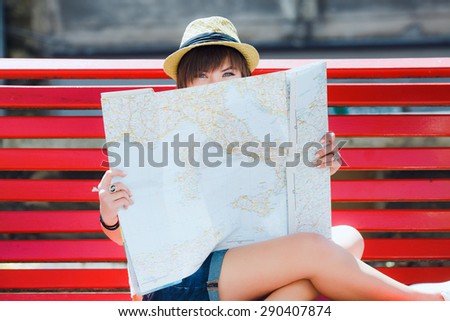 Pretty tourist young woman with sly glance, sitting on red bench and hiding on the map of Europe. With shortcut dark-blonde hair. Wearing stylish summer hat and denim shorts. Looking at camera.