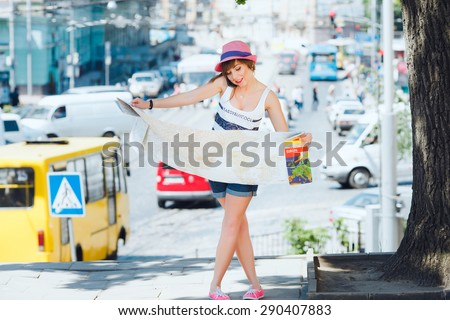 Pretty tourist girl speaking on the mobile phone and holding a big map of Europe. On traffic background. With shortcut dark-blonde hair. Wearing stylish pink hat and denim shorts.