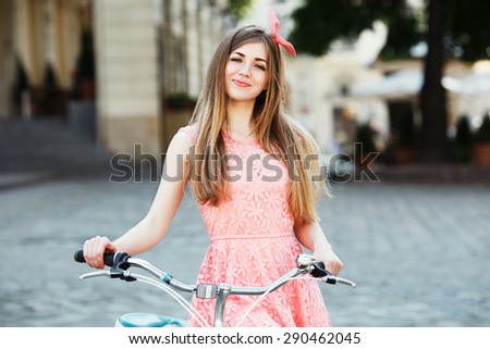 Pretty blond-brown girl holding bicycle. Wearing pink head wrap and dress with a pattern of flowers. Looking at the camera. In the old European city. Close up.