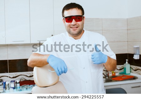 Dentist in red protective glasses showing thumbs up, over medical office background.