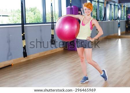 Young beautiful redhead woman is standing near the mirror with fitball in gym, full length